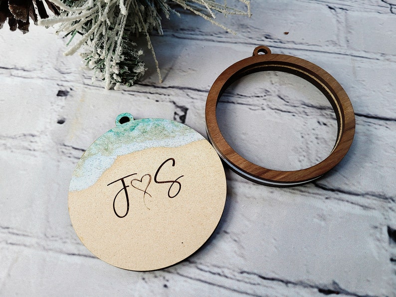 May include: A wooden ornament with a light blue and white watercolor design resembling a beach scene. The ornament has the initials "J & S" with a heart symbol in between. The ornament is attached to a wooden hoop.