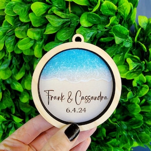 May include: A round wooden ornament with a blue and white beach scene inside. The ornament is personalized with the text "Frank & Cassandra" and the date "6.4.24".