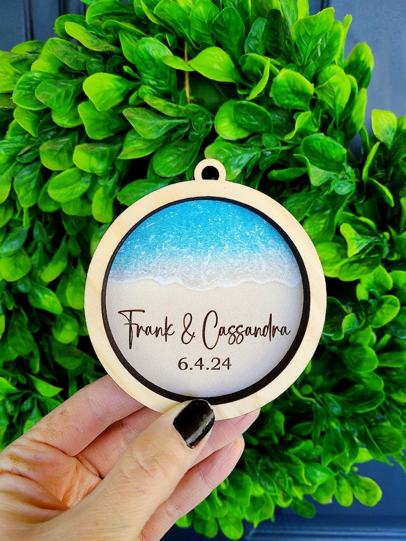 May include: A round wooden ornament with a blue and white beach scene inside. The ornament is personalized with the text "Frank & Cassandra" and the date "6.4.24".