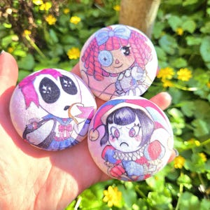 May include: Three circular buttons with cartoon illustrations. One depicts a doll with pink hair and a monocle, another a sad jester, and the third a skeleton-like character. The buttons are held in a hand against a backdrop of green leaves and yellow flowers.