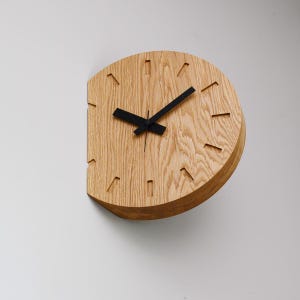 Könnte beinhalten: Eine halbkreisförmige Wanduhr aus Holz mit hellbraunem Finish. Die Uhr hat schwarze Zeiger und Stundenmarkierungen. Die Uhr ist an einer weißen Wand befestigt.