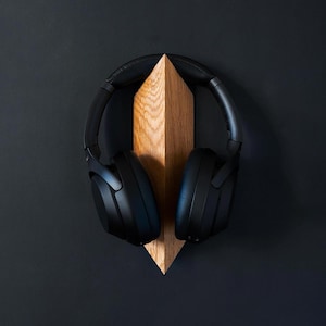 Peut inclure: Casque audio noir supra-auriculaire suspendu à un support en bois. Le support est en forme de triangle avec un point en haut.