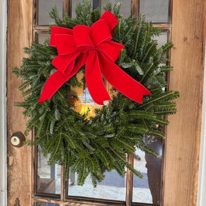 May include: A fresh evergreen wreath with a vibrant red velvet bow hangs on a wooden door. The wreath is made of lush green pine branches, creating a festive holiday decoration. The door has a glass window and a brass doorknob.