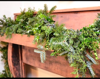 Puede incluir: Una repisa de madera rústica adornada con una guirnalda de vegetación fresca. La guirnalda presenta una mezcla de ramas de hoja perenne, boj y bayas, creando una exhibición natural y festiva. La madera tiene un tono marrón cálido.
