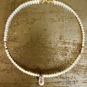 May include: A delicate necklace featuring small, white pearls and a gold clasp. The necklace has a teardrop-shaped pendant with pink and purple hues. The jewelry is displayed on a gold-toned surface.