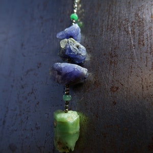 May include: A necklace featuring a silver chain with a series of natural gemstones. The necklace includes rough-cut, blue-purple stones, green stones, and small beads. The pendant has a gold-colored metal base with a red bead.
