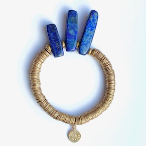 Könnte beinhalten: Ein goldfarbenes Armband mit einem kreisförmigen Design aus kleinen, gestapelten Metallringen. Oben sind drei rechteckige, tiefblaue Lapislazuli-Steine angebracht. Ein kleiner, runder Goldanhänger mit Text hängt am Armband.