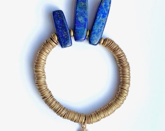 May include: A gold-coloured bracelet with a circular design made of small, stacked metal rings. Three rectangular, deep blue lapis lazuli stones are set at the top. A small, round gold charm with text hangs from the bracelet.