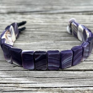 May include: A bracelet composed of rectangular, polished beads featuring alternating bands of deep purple and white. The beads are strung together in a curved line, and the ends of the bracelet are secured with black cord ties that are visible at one side.