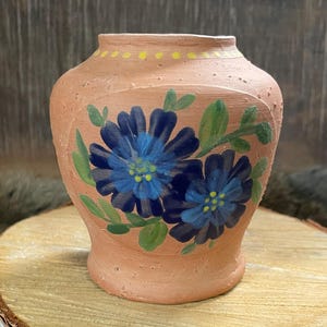 May include: A terracotta vase featuring hand-painted blue flowers and green leaves. The vase has a rounded body and a narrow neck, with yellow dots around the rim. It rests on a wooden slice.