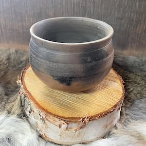 May include: A handcrafted ceramic bowl with a rounded form and a broad opening. The bowl displays a mix of grey and brown hues, with darker patches near the base. It rests on a natural wood slice, highlighting the rustic texture of the pottery.