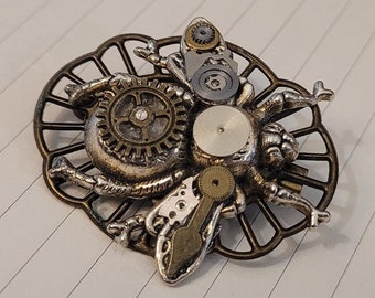 May include: A silver and bronze steampunk-style insect brooch with intricate gears and a detailed design. The brooch is set on a decorative, openwork frame.