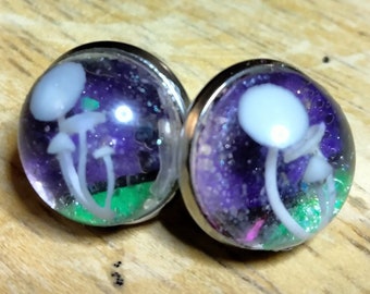 May include: A pair of silver stud earrings with a clear resin dome containing a purple and green glitter background with white mushroom silhouettes.