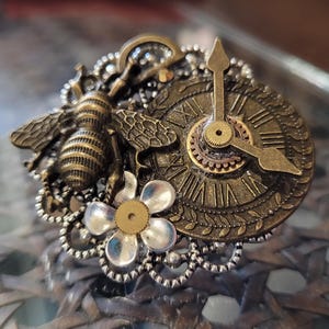 May include: A steampunk-style brooch featuring a detailed bee, a clock face with Roman numerals, and a flower. The brooch is made of bronze and silver-toned metal, with intricate filigree and gear details.