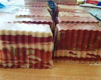 May include: A close-up of several bars of handmade soap. The soap is layered with shades of brown, white, and green, creating a striped pattern. The soap is arranged on a wooden surface.