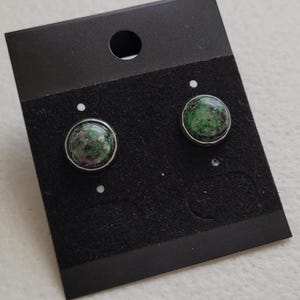 Może przedstawiać: Para okrągłych kolczyków sztyftów ze srebrną oprawą. Każdy kolczyk ma zielony i czerwony nakrapiany kamień. Kolczyki są prezentowane na czarnej karcie do kolczyków z okrągłym otworem u góry.