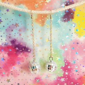 May include: A pair of silver disco ball earrings hanging from gold chains against a colorful background with stars.