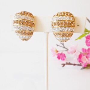 May include: A pair of gold-toned clip-on earrings. Each earring is oval-shaped and covered in alternating rows of clear and amber-colored rhinestones. The earrings are displayed on a white stand, with pink flowers in the background.