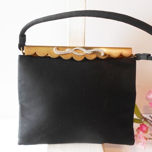 May include: A black satin evening bag with a gold metal clasp featuring a rhinestone design. The bag has a single top handle.
