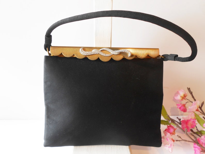 May include: A black satin evening bag with a gold metal clasp featuring a rhinestone design. The bag has a single top handle.