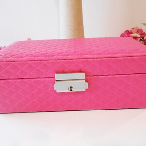 May include: A rectangular, hot pink jewelry box with a quilted pattern. The box has a silver-colored clasp and a keyhole. The background includes pink flowers and green leaves.