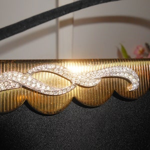 May include: A gold-colored clutch purse with a scalloped edge and a rhinestone design. The rhinestone design is a wavy line with two loops.