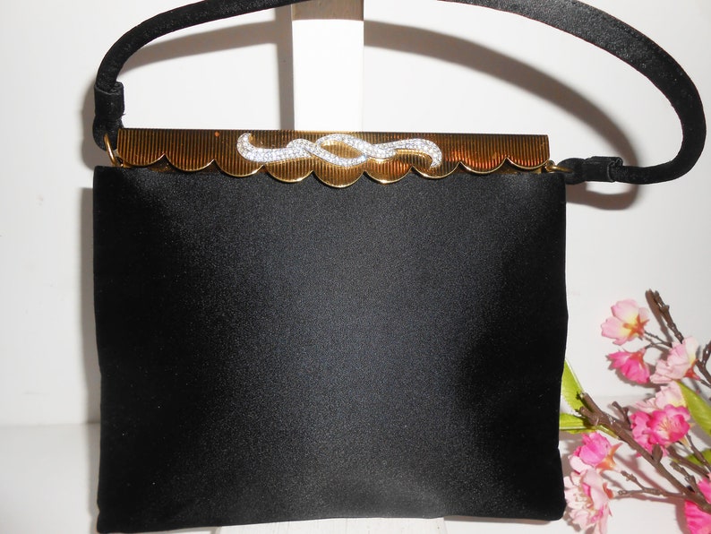 May include: A black satin evening bag with a gold clasp and a rhinestone design. The bag has a short, black handle.