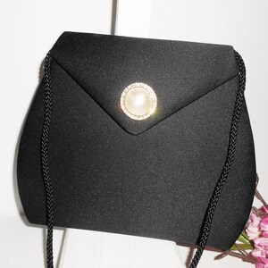 May include: A black satin evening bag with a pearl and rhinestone embellishment. The bag has a long black chain strap.