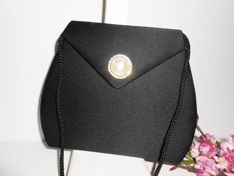 May include: A black satin evening bag with a pearl and rhinestone embellishment. The bag has a long black chain strap.