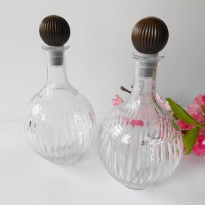 May include: Two clear glass bottles with ribbed designs and brown wooden stoppers. The bottles are empty and have a round shape.