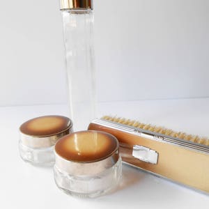 May include: A set of vintage bathroom accessories in a mid-century modern style. The set includes a glass bottle with a gold lid, two glass jars with gold lids and a brush with a brown handle and silver trim.