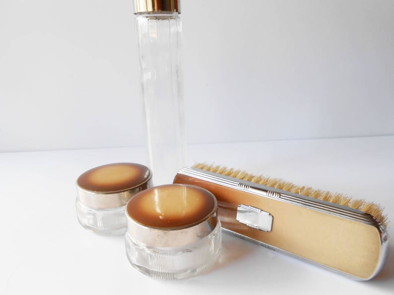 May include: A set of vintage bathroom accessories in a mid-century modern style. The set includes a glass bottle with a gold lid, two glass jars with gold lids and a brush with a brown handle and silver trim.