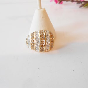 May include: A gold-toned ring with a rounded, oval shape. The ring is encrusted with alternating rows of clear and amber-colored crystals. The ring is displayed on a white stand, with a soft, blurred background.