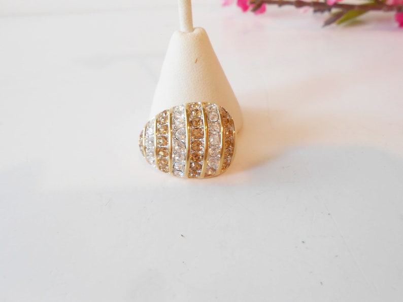 May include: A gold-toned ring with a rounded, oval shape. The ring is encrusted with alternating rows of clear and amber-colored crystals. The ring is displayed on a white stand, with a soft, blurred background.