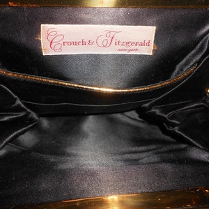 May include: Inside view of a black satin clutch purse with a gold metal frame. The purse has a small pocket on the inside and a label that reads "Crouch & Fitzgerald, New York".