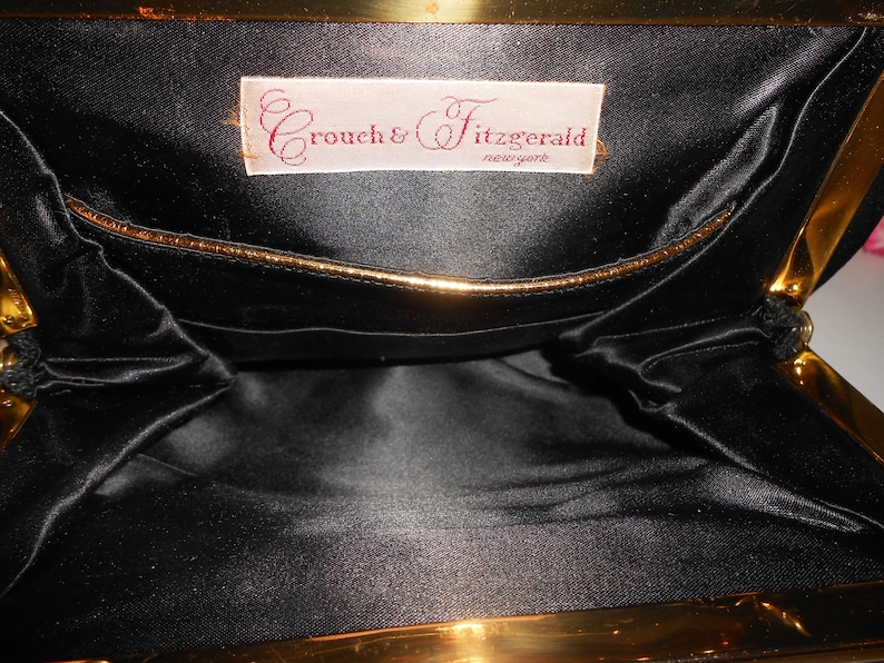May include: Inside view of a black satin clutch purse with a gold metal frame. The purse has a small pocket on the inside and a label that reads "Crouch & Fitzgerald, New York".