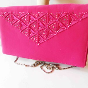 May include: A hot pink satin clutch with a silver chain strap. The clutch has a geometric design with rhinestones on the front.