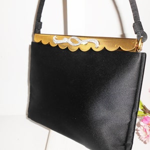 May include: A black satin evening bag with a gold metal clasp featuring a rhinestone design. The bag has a short strap.