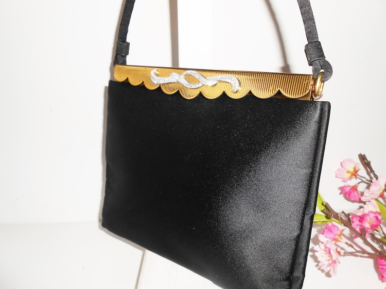May include: A black satin evening bag with a gold metal clasp featuring a rhinestone design. The bag has a short strap.