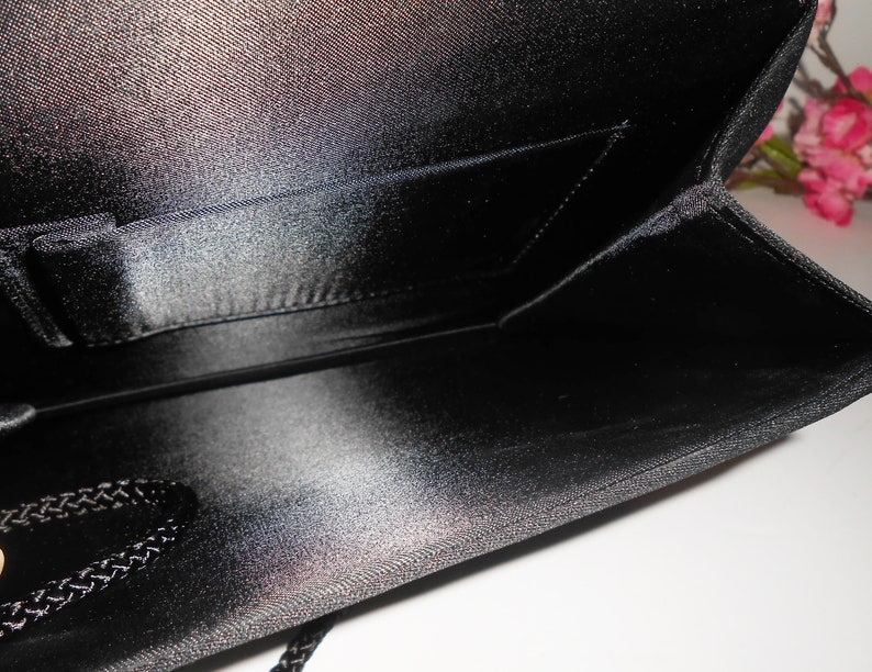 May include: Black satin clutch bag with a small interior pocket. The bag has a zipper closure and a black braided cord.