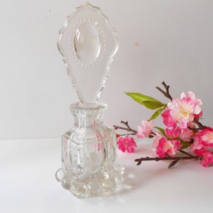 May include: A clear glass perfume bottle with a decorative, ornate stopper. The bottle has a square base with a textured design and a round, bulbous bottom. The stopper is shaped like a teardrop with a scalloped edge and a raised, oval design in the center.