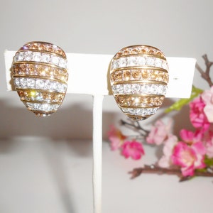 May include: A pair of oval-shaped clip-on earrings. The earrings feature alternating horizontal bands of clear and amber-colored crystals set in a gold-tone metal. The earrings are displayed on a white stand against a soft background with pink flowers.