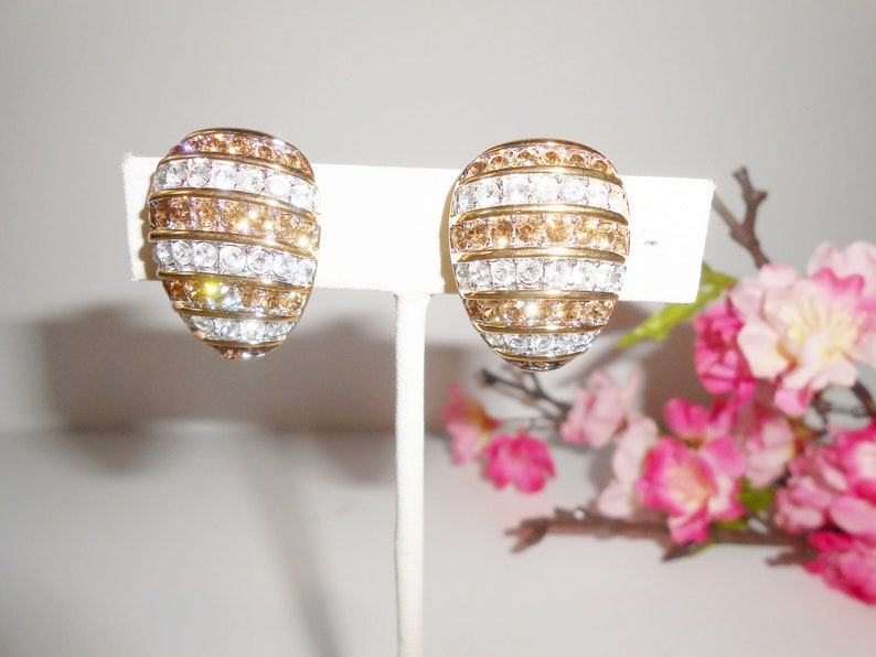 May include: A pair of oval-shaped clip-on earrings. The earrings feature alternating horizontal bands of clear and amber-colored crystals set in a gold-tone metal. The earrings are displayed on a white stand against a soft background with pink flowers.
