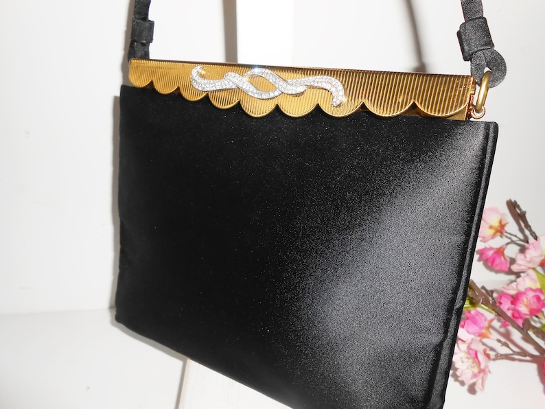 May include: A black satin evening bag with a gold metal frame and a rhinestone embellishment. The bag has a scalloped edge and a chain strap.