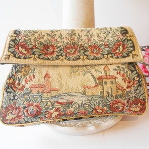 May include: A vintage tapestry clutch bag with a floral design and a scene of a small town with buildings and trees. The bag is beige with red, green, and brown accents.