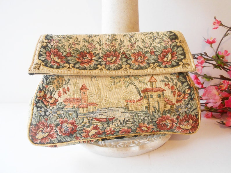 May include: A vintage tapestry clutch bag with a floral design and a scene of a small town with buildings and trees. The bag is beige with red, green, and brown accents.