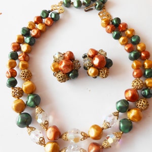 May include: A two-strand necklace and matching earrings with green, gold, and orange beads. The necklace has a gold clasp and the earrings are made of a cluster of beads.