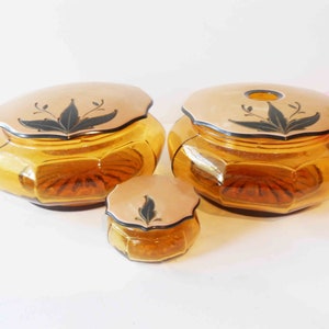 May include: Set of three amber glass vanity jars with lids. The lids are cream colored with black floral designs.
