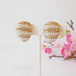 May include: A pair of gold-toned clip-on earrings. Each earring has an oval shape and is adorned with alternating rows of clear and amber-colored rhinestones. The earrings are displayed on a white stand against a soft background.