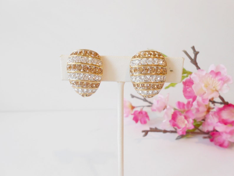 May include: A pair of gold-toned clip-on earrings. Each earring has an oval shape and is adorned with alternating rows of clear and amber-colored rhinestones. The earrings are displayed on a white stand against a soft background.